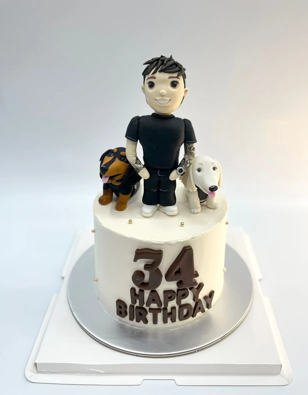 White 34th birthday cake topped with a fondant man figurine and his two dogs, chocolate Happy Birthday lettering