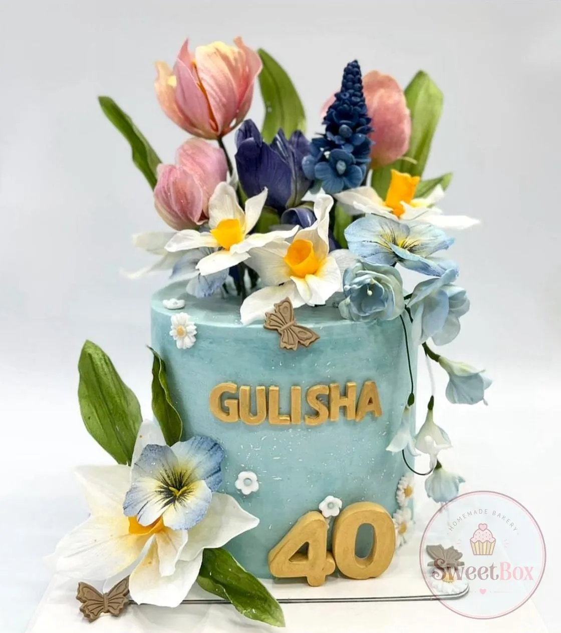Tall light blue 40th birthday cake with sugar tulips, daffodils and pansies, gold name lettering and a gold number 40