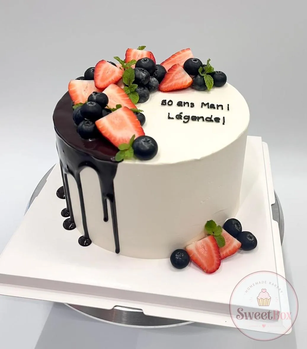White cake with dark chocolate drip, strawberries and blueberries and a short French message piped in black