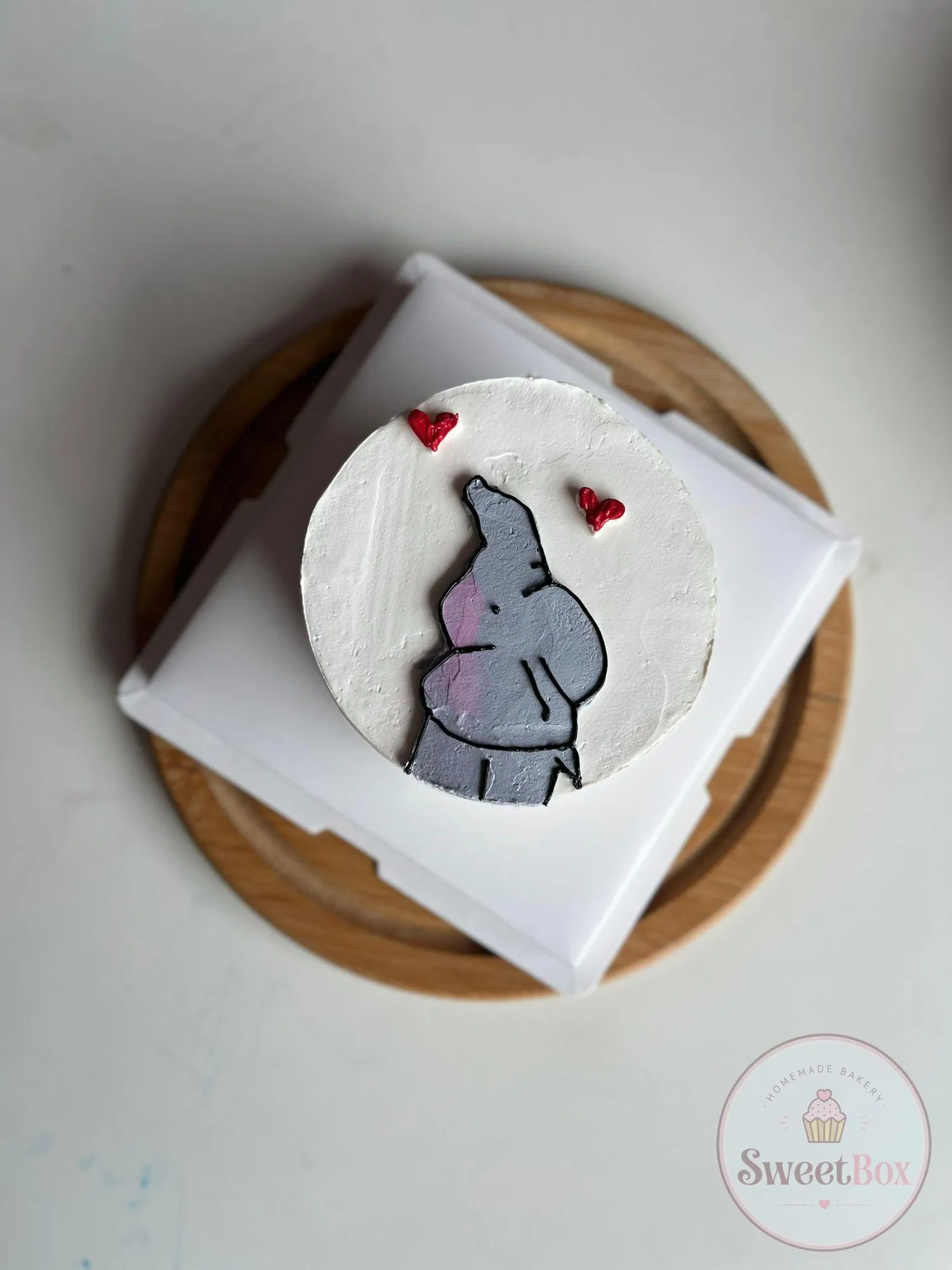 White bento cake with a hand-piped gray baby elephant and two red hearts, on a white box and wooden board