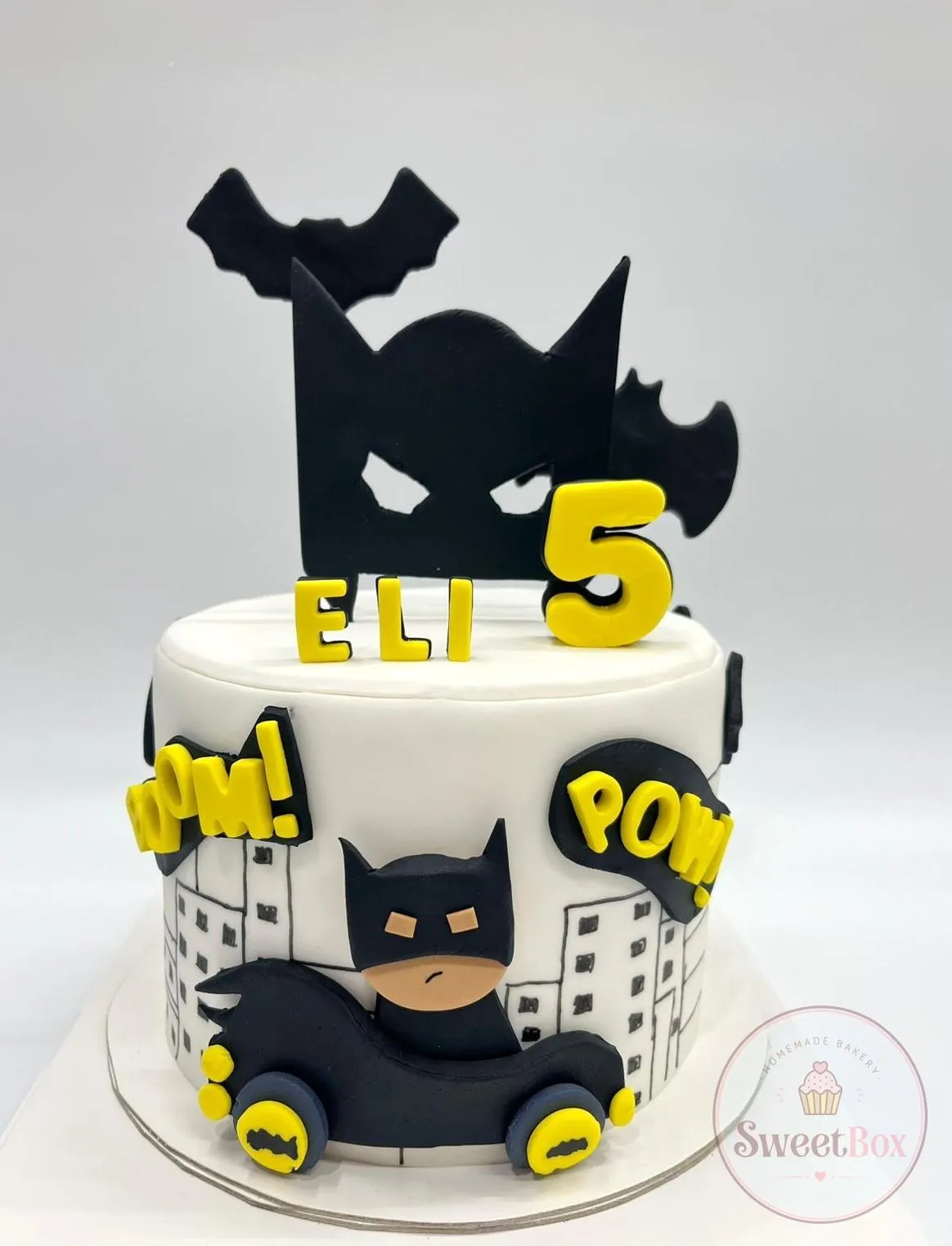 White Batman cake with a black mask topper, drawn city skyline, Batmobile, BOOM and POW bubbles and a yellow name and number 5