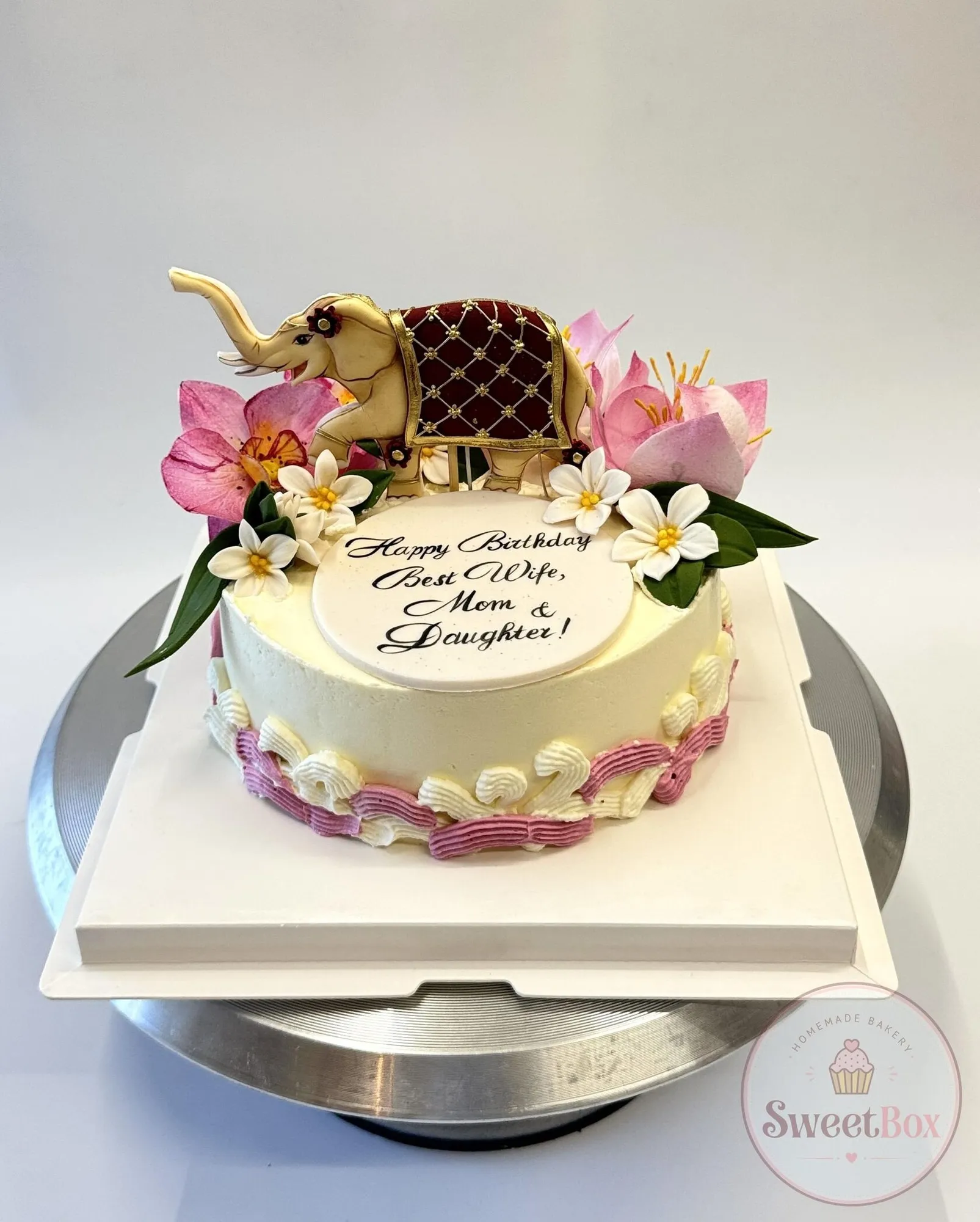 Cream birthday cake with a gold Thai elephant topper, pink lotus, frangipani flowers and a piped message