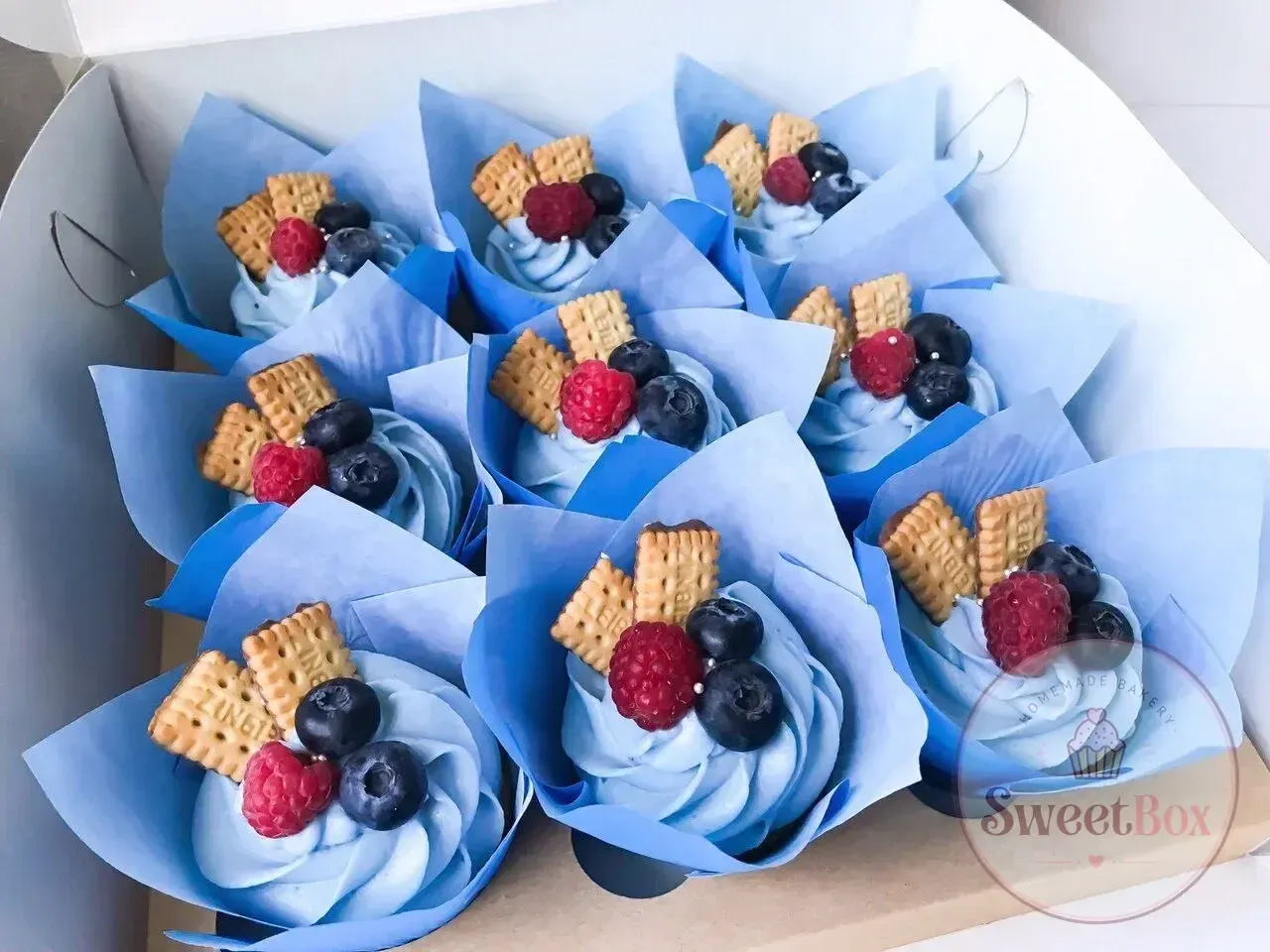 Box of blue cream cupcakes topped with raspberries, blueberries and biscuit pieces