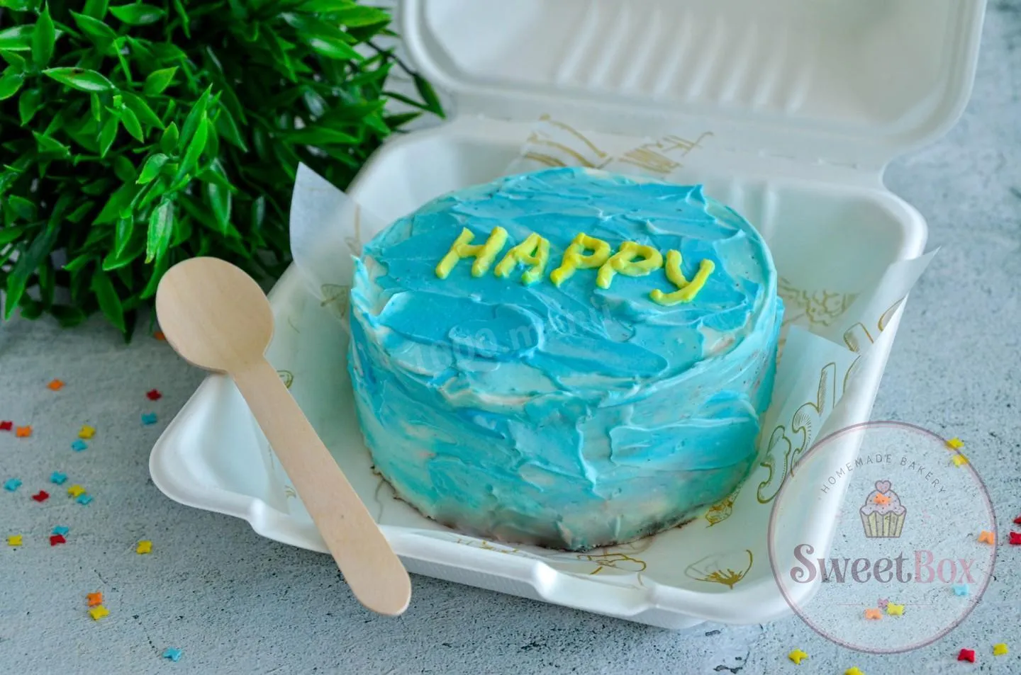 Blue textured bento cake with yellow Happy lettering in a takeout box with a wooden spoon