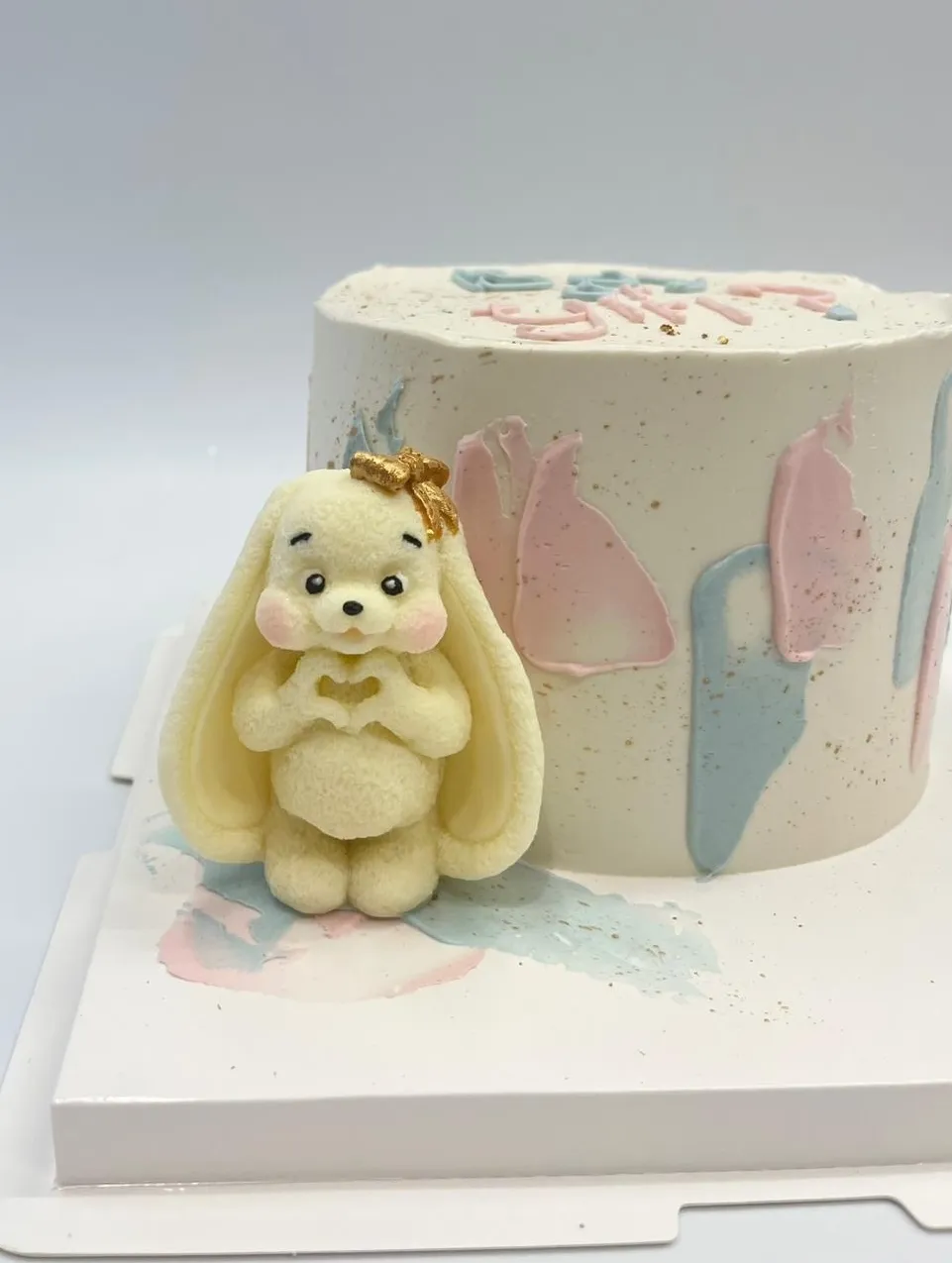 White cake with pink and blue brushstrokes and a cream bunny figure holding a heart