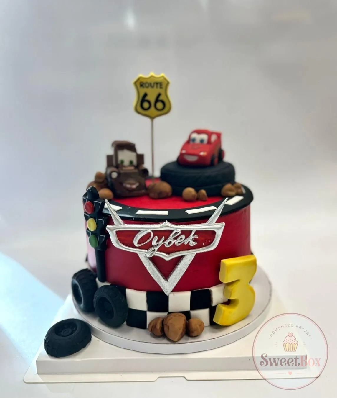 Red cake with a race-car and tow-truck figures, Route 66 sign, tires, traffic light, checkered base and a yellow number 3