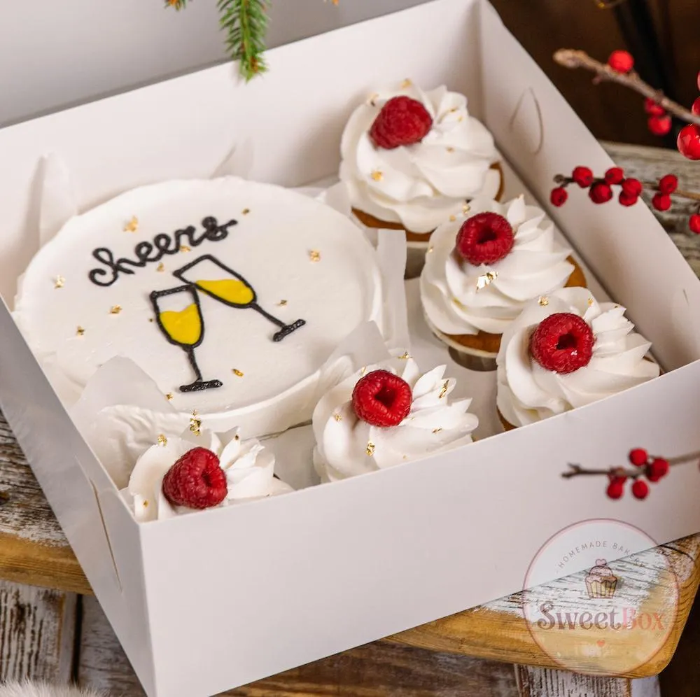 Cheers bento cake with a champagne glasses drawing and cupcakes topped with raspberries in a box