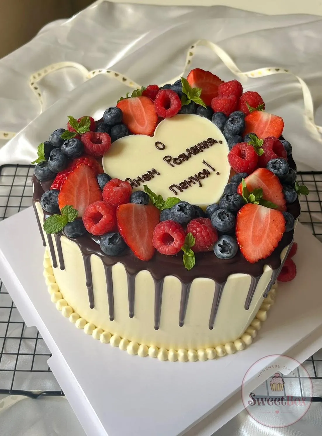 Cream cake with dark chocolate drip, a wreath of strawberries, raspberries, blueberries and mint and a white chocolate heart plaque