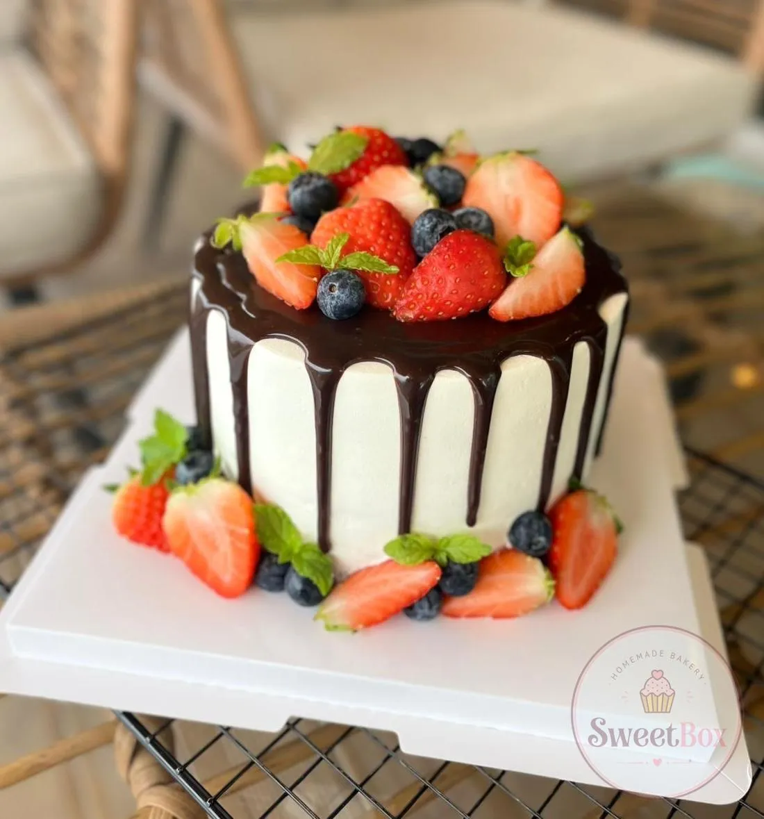 White cake with dark chocolate drip crowned with fresh strawberries, blueberries and mint, more berries around the base