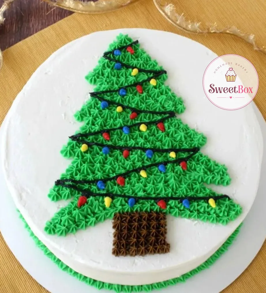 White round cake with a green piped Christmas tree, a black string of colorful lights and a green piped border