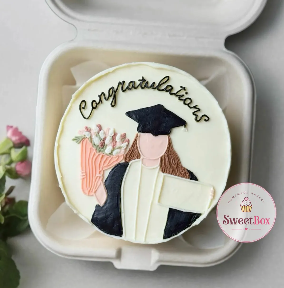 Bento cake with Congratulations script and a piped graduate in a black gown holding a bouquet and a diploma