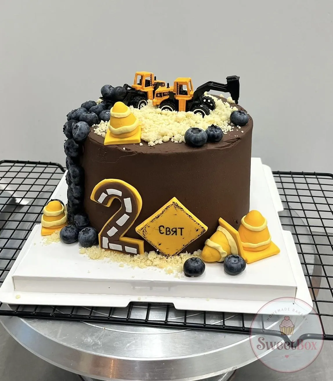 Chocolate cake with toy diggers on crumble, yellow traffic cones, blueberries, a road-pattern number 2 and a name sign