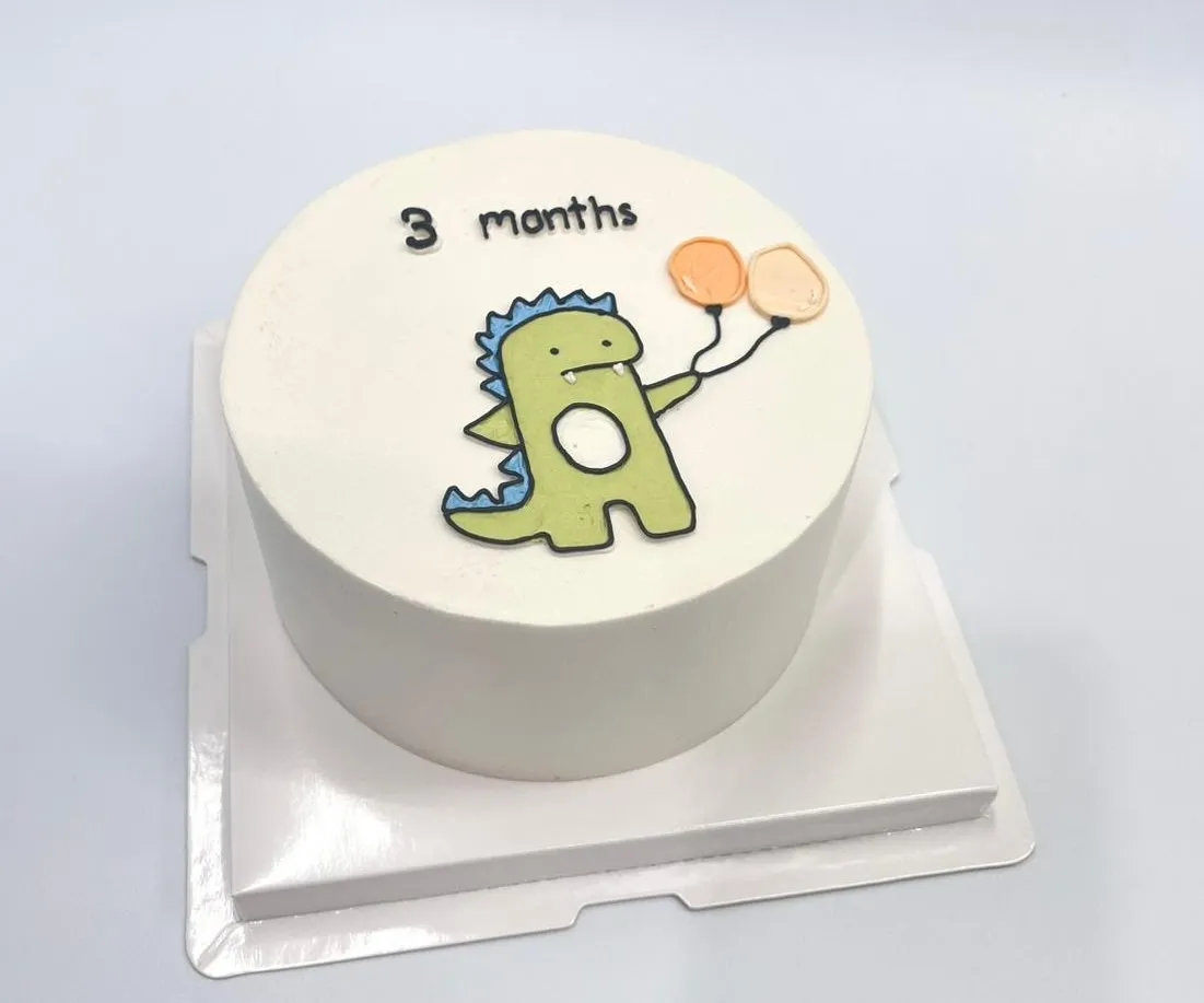White bento cake with a hand-drawn green dinosaur holding two orange balloons and 3 months lettering