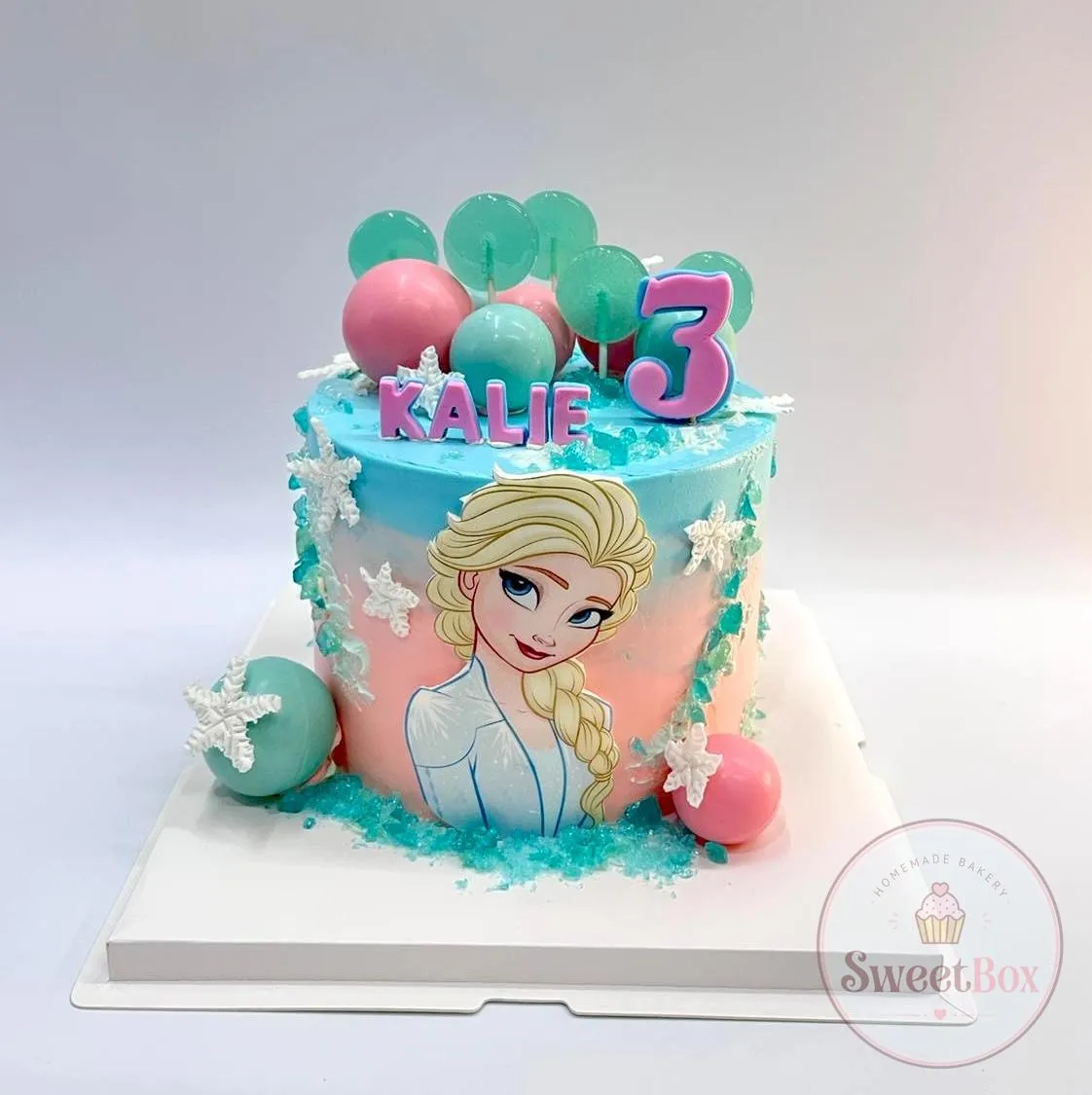 Pink and blue Frozen-themed cake with an Elsa picture, snowflakes, spheres, a number 3 and a child's name