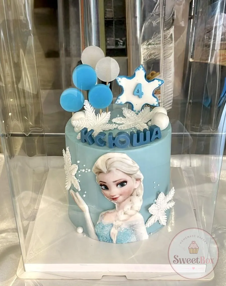 Pale blue Frozen-themed cake with an Elsa picture, white snowflakes, lollipops and a number 4 in a clear box