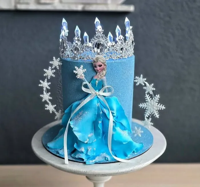Tall blue Frozen cake with an Elsa ball gown, silver crystal crown and snowflakes on a cake stand