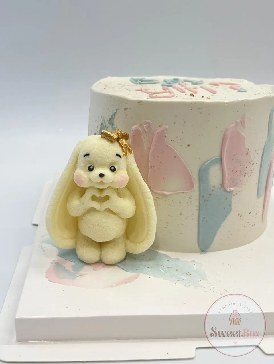 White cake with pink and blue brushstrokes, gold flecks and a white chocolate bunny figure making a heart with its paws