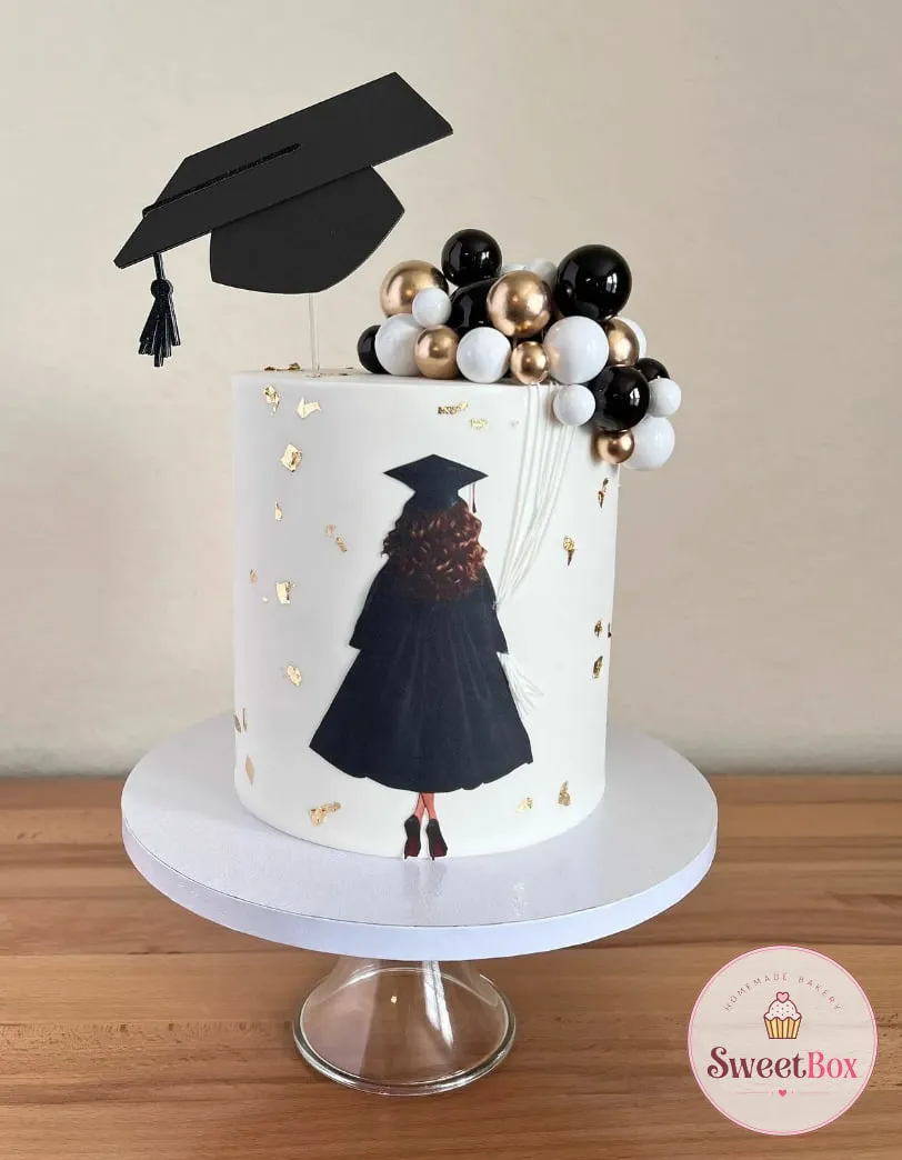 Tall white graduation cake with a painted graduate in a black gown, gold leaf, a mortarboard topper and black, white and gold balls