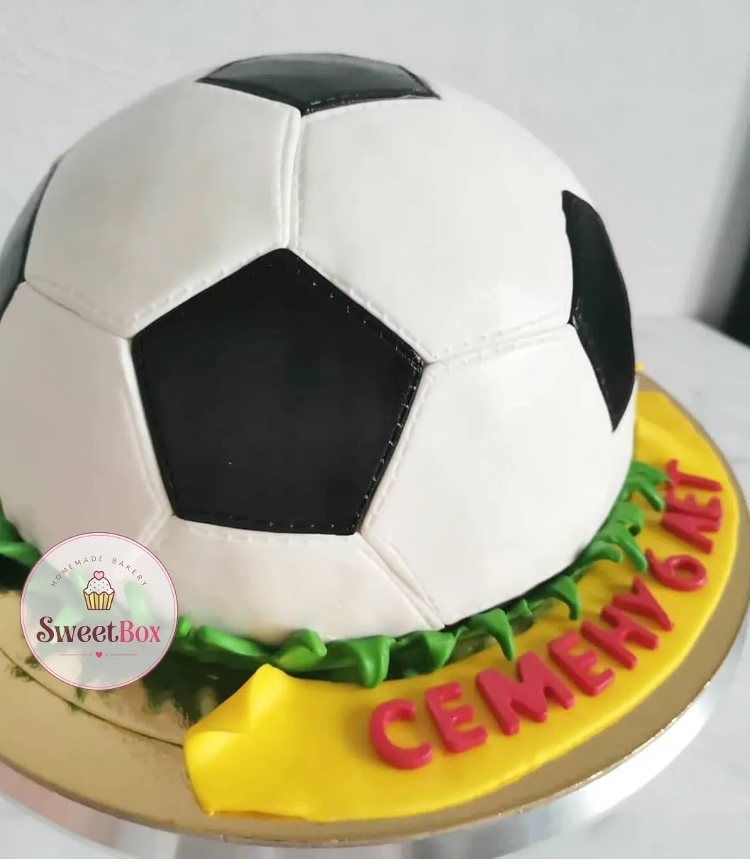 Dome cake shaped like a soccer ball on piped green grass, with a yellow ribbon carrying a child’s name and age in red Russian letters