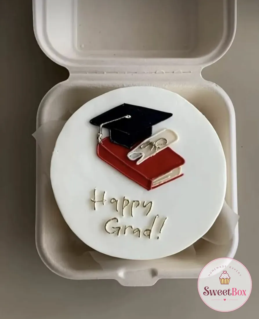 White bento cake in a takeout box with a piped mortarboard, red book and diploma and gold Happy Grad! lettering