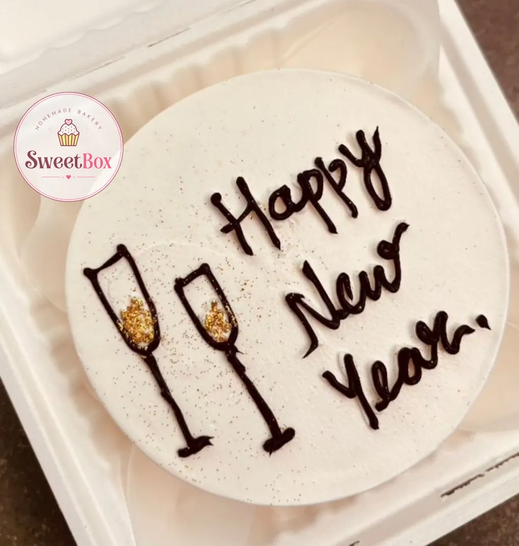 White bento cake in a takeout box with piped Happy New Year lettering and two champagne glasses filled with gold glitter