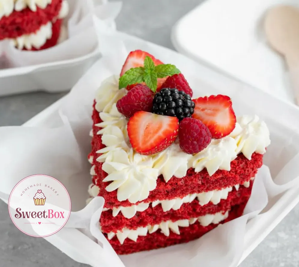 Small heart-shaped red velvet layer cake with piped cream, strawberries, raspberries, a blackberry and mint in a box