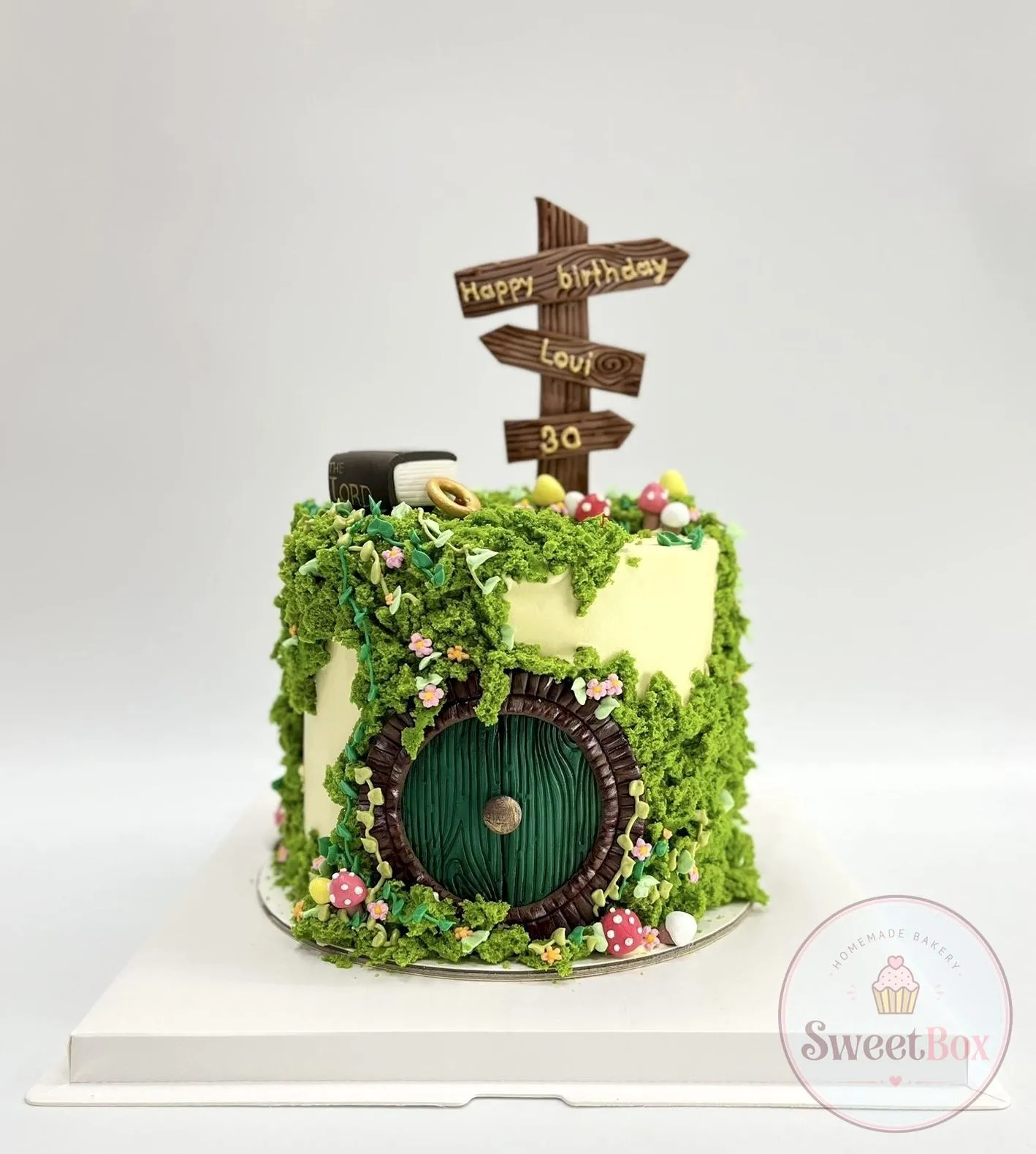 Cake covered in green sponge moss with a round green fairy-tale door, tiny mushrooms, a book, a gold ring and a wooden signpost