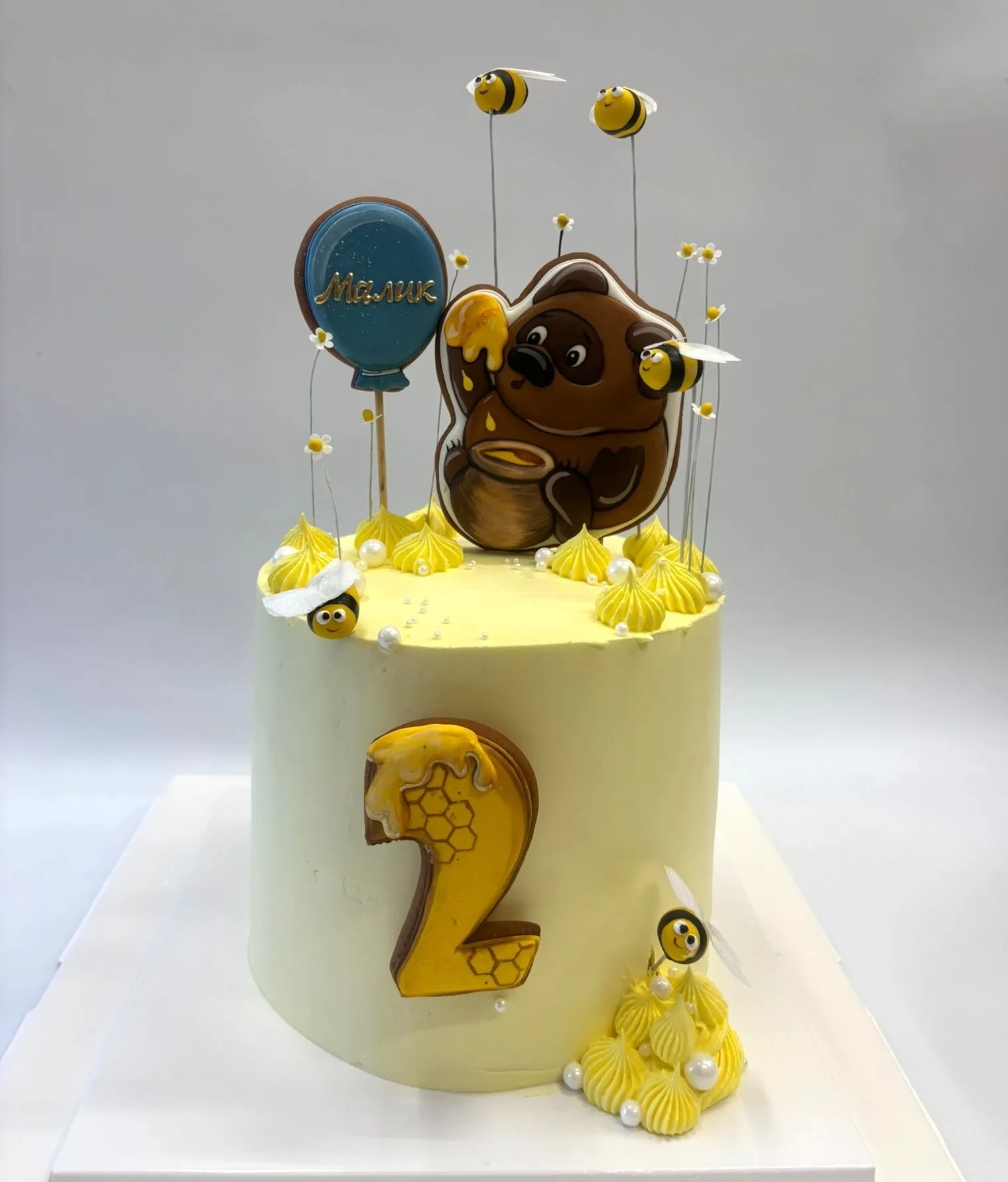 Pale yellow second birthday cake with a honey bear cookie topper, flying bees and a gold number 2