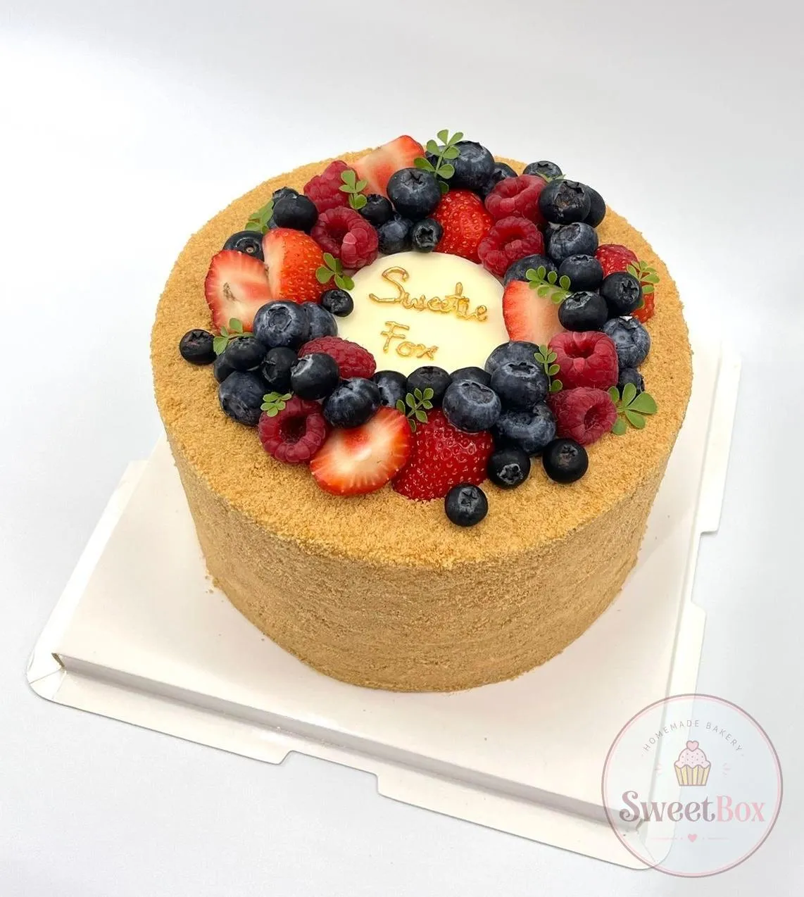 Crumb-coated honey cake with a wreath of strawberries, raspberries and blueberries around a white plaque with gold Sweet Box lettering