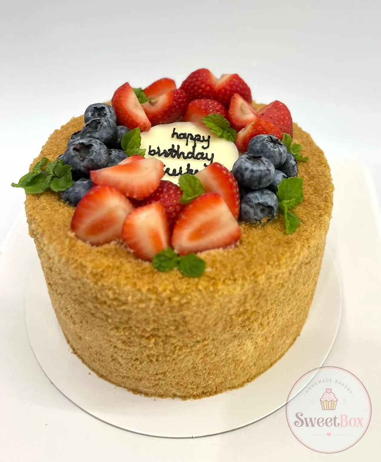 Tall crumb-coated honey cake topped with strawberries, blueberries, mint and a white happy birthday plaque
