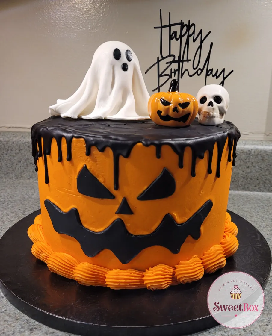 Orange jack-o’-lantern cake with a black drip, a white ghost, a small pumpkin, a skull and a Happy Birthday topper