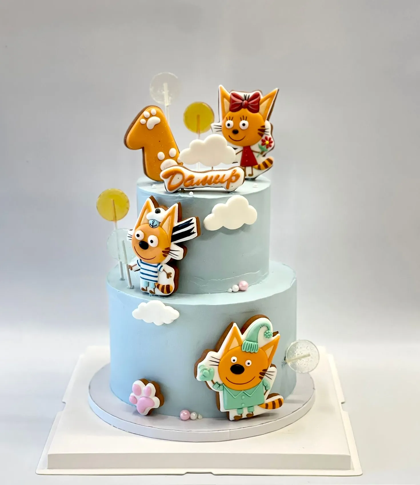 Two-tier light blue first birthday cake decorated with Kid-E-Cats cookies, clouds and a name topper