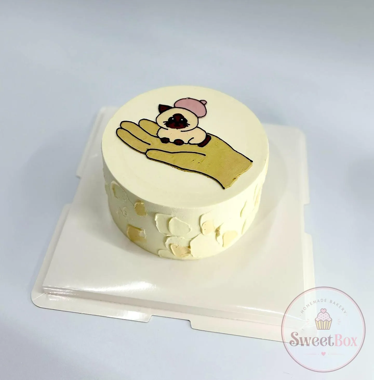 Cream bento cake with a drawing of a tiny kitten in a pink hat sitting in an open hand