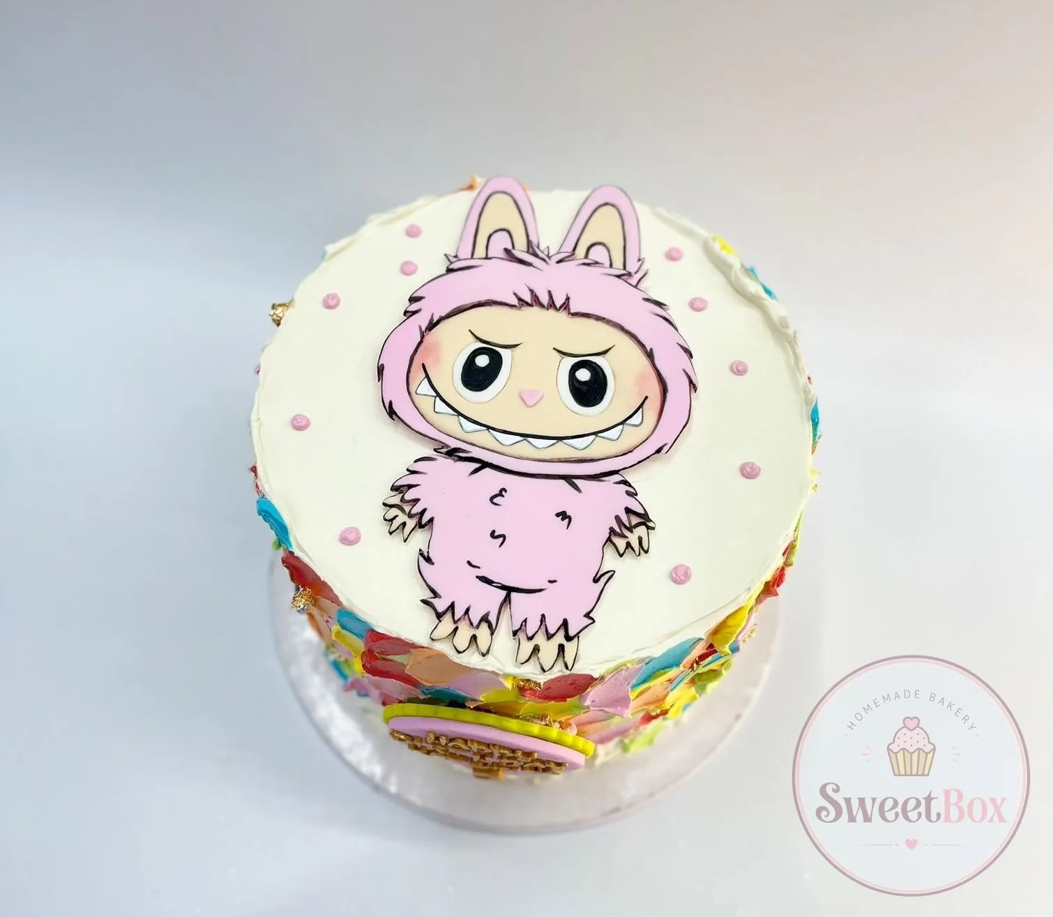 White cake seen from above with a pink furry monster character drawing, pink dots and rainbow buttercream sides