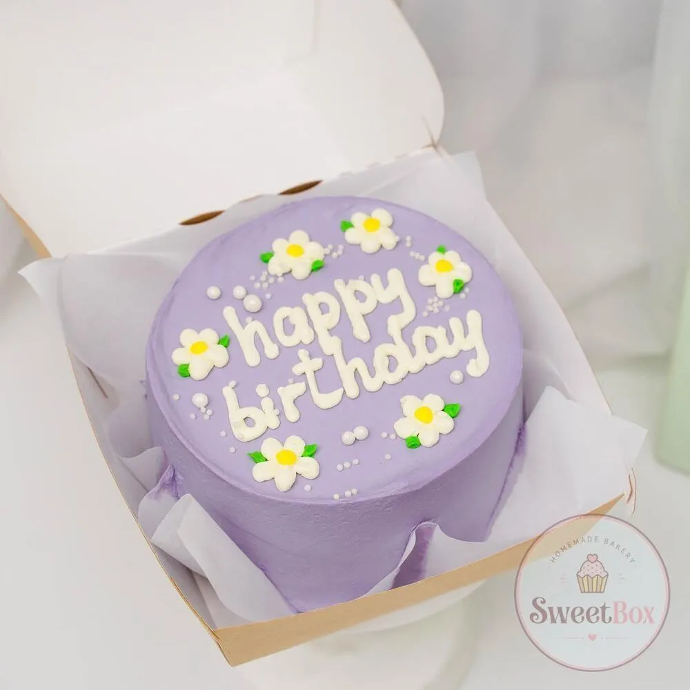 Lilac bento cake with white piped happy birthday lettering, white daisies and sugar pearls in an open kraft box
