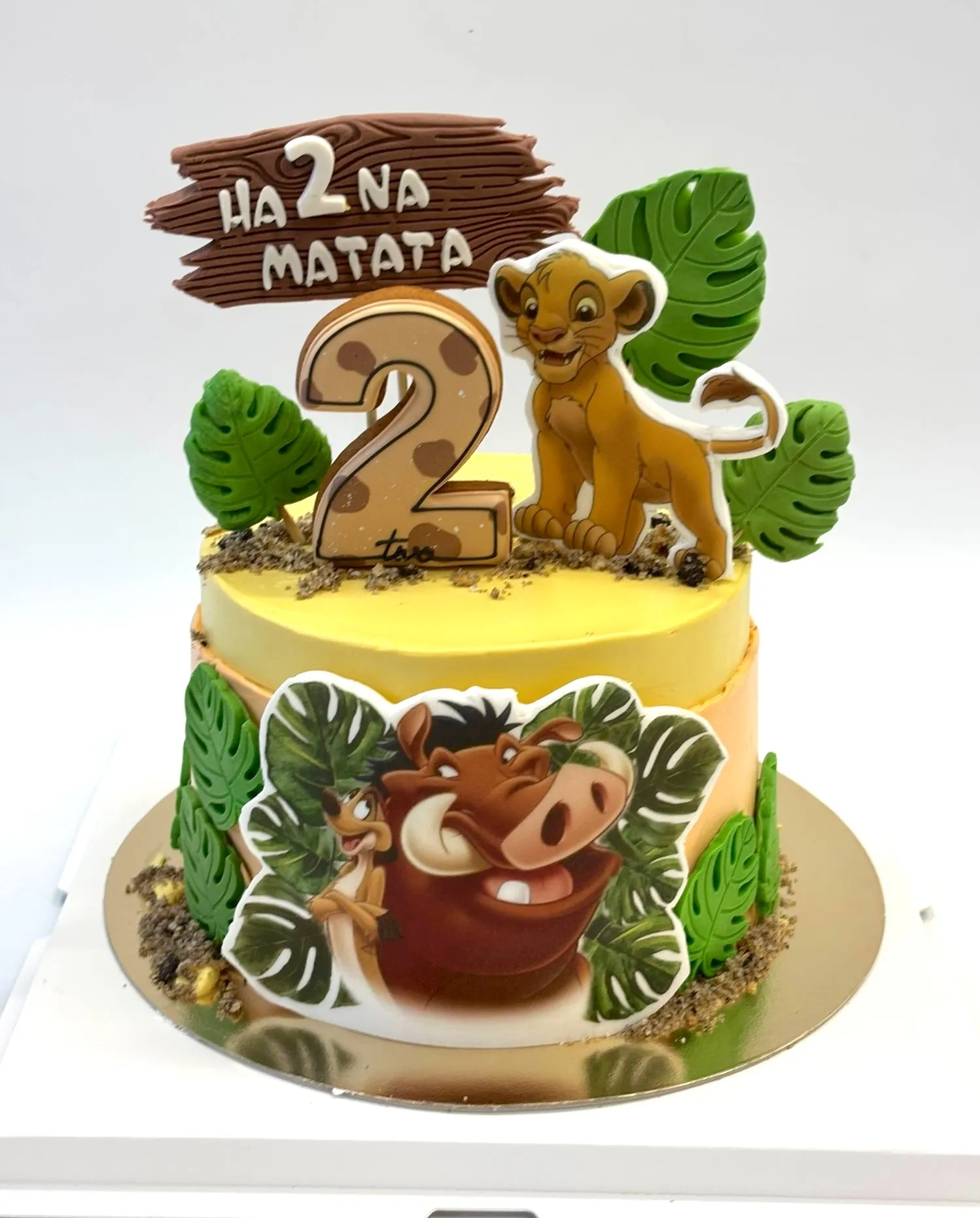 Yellow second birthday cake with a lion cub topper, warthog and meerkat print, green tropical leaves and a Ha-2-na Matata sign