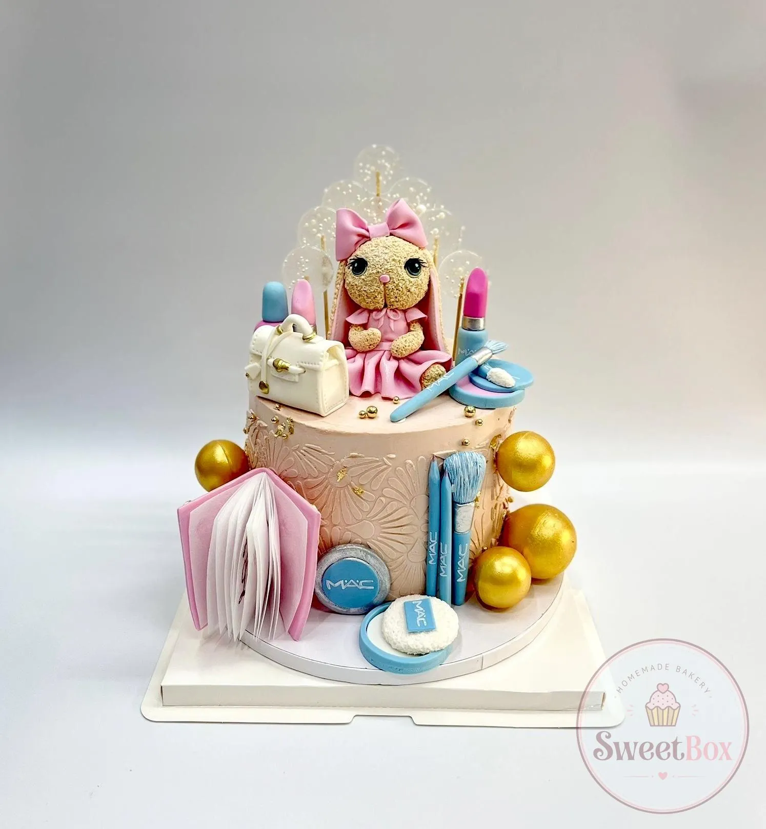 Peach cake with a plush-style bunny in a pink dress and bow, a white handbag, lipsticks, brushes, a pink book and gold spheres