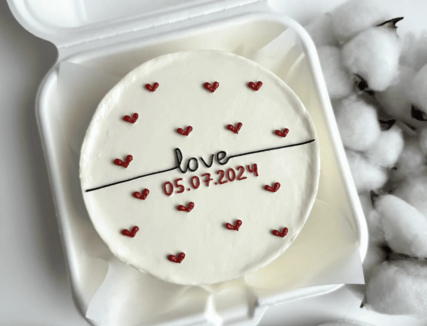 White bento cake with love lettering, a date and tiny red piped hearts in a takeaway box