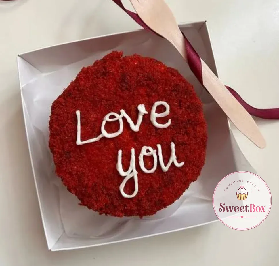 Red velvet bento cake covered in red crumbs with white Love you lettering in a white box with a wooden fork
