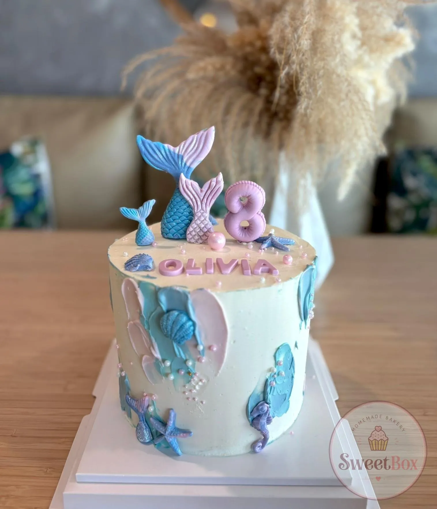 White cake with blue and pink brushstrokes, glittery mermaid tail toppers, seashells, a seahorse, an Olivia name and a pink number 8