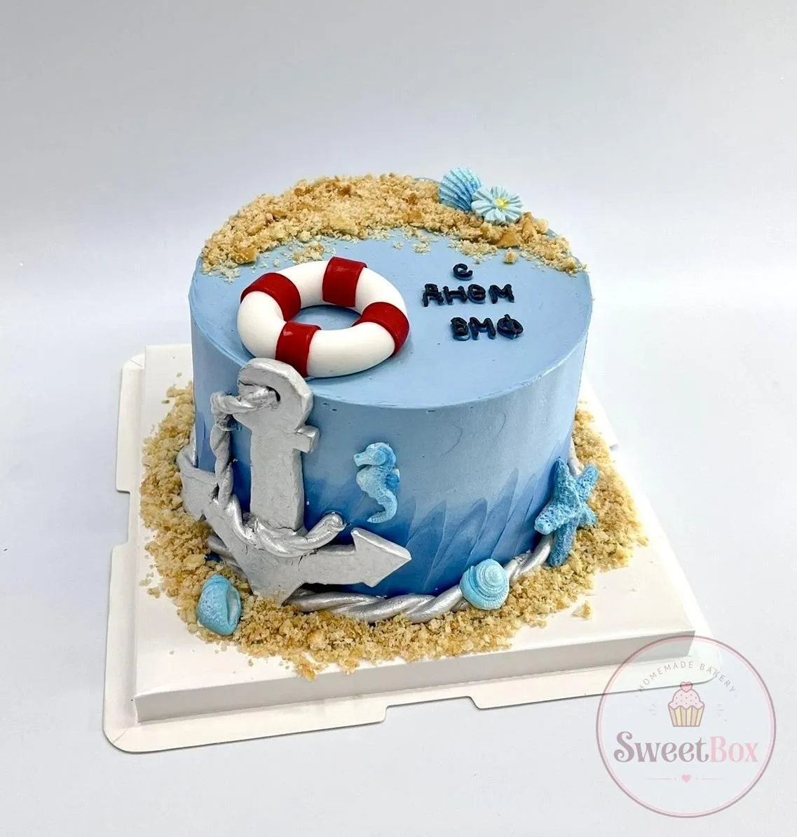 Blue nautical cake with a silver anchor, a red and white life ring, seashells and a sandy crumb border