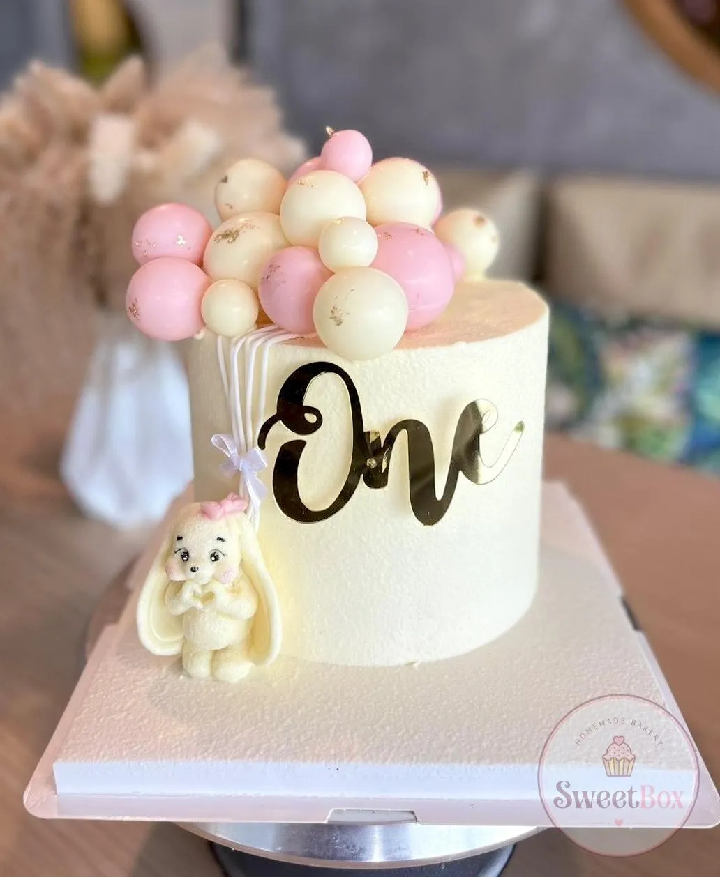 White first birthday cake with a pink and white balloon cluster, gold One topper and a bunny