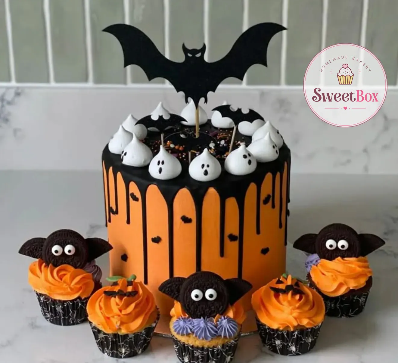 Orange cake with a black drip, meringue ghosts and a black bat topper, with Oreo bat and pumpkin cupcakes in front