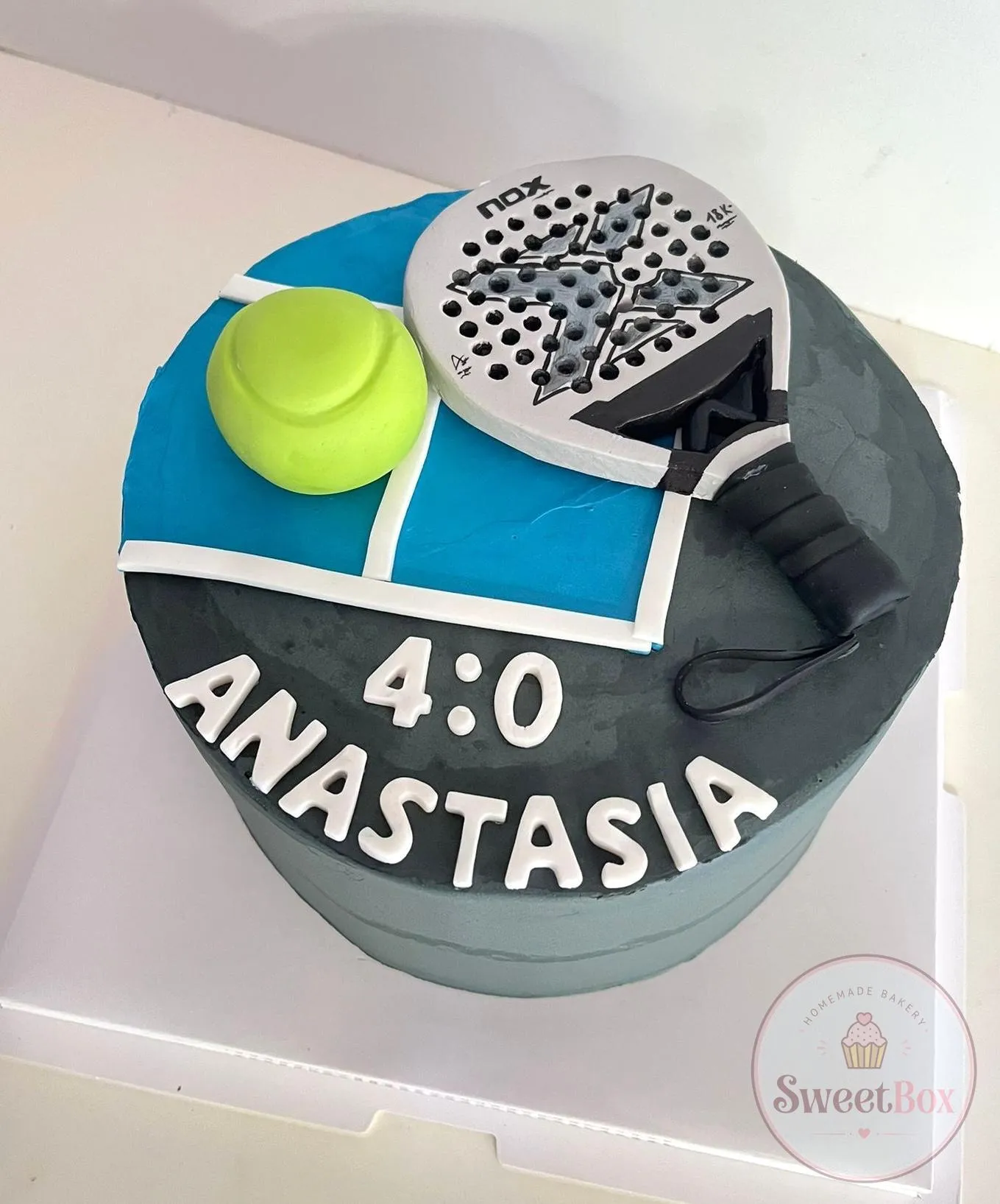 Gray padel cake with a blue court on top, a white padel racket, a yellow ball, the score 4:0 and a name in white letters