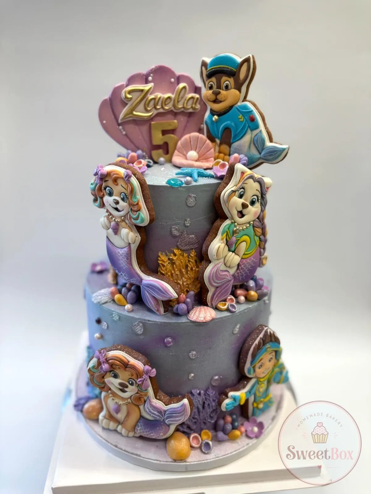 Two-tier lilac cake with puppy cookies in mermaid tails, coral, shells and a seashell name topper with gold number 5