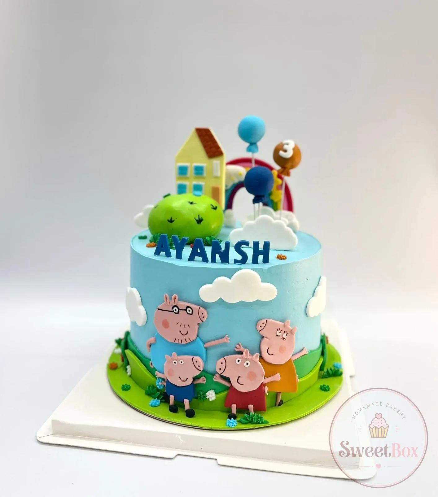 Sky blue cake with a cartoon pig family, clouds, a yellow house, rainbow, balloons, green hills and an Ayansh name