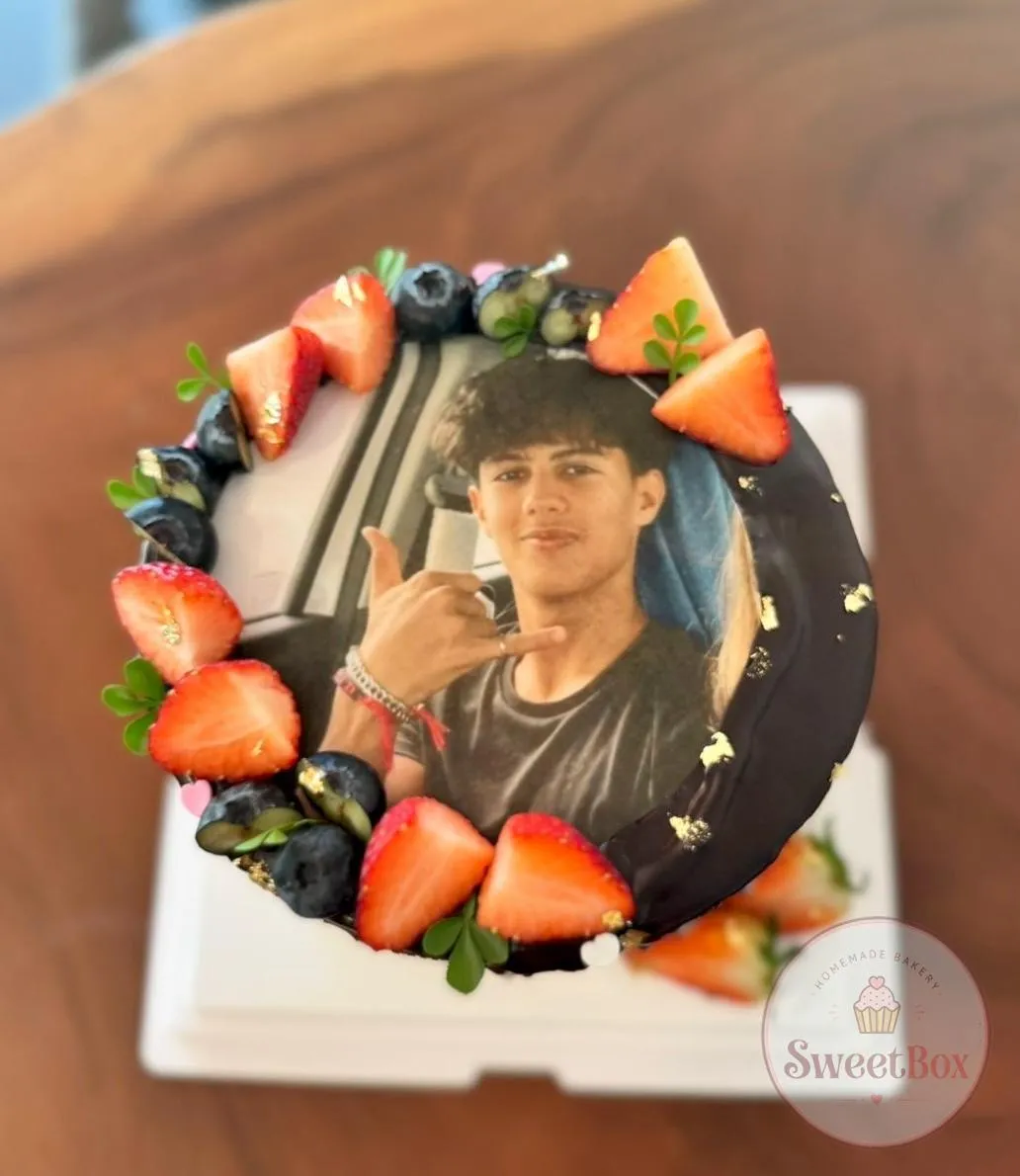 Top view of a cake with an edible portrait of a smiling teen framed by strawberries, blueberries, gold leaf and chocolate glaze
