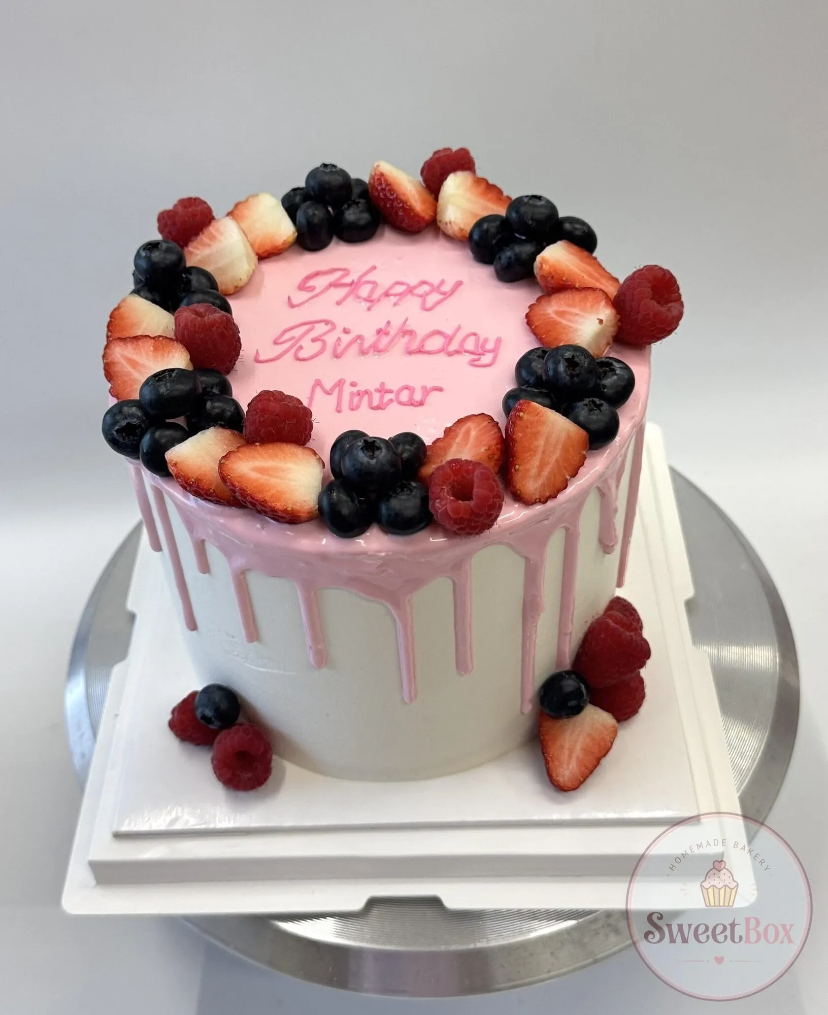 White cake with pink drip, a ring of strawberries, raspberries and blueberries and a pink Happy Birthday Mintar message