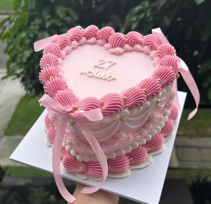Pink vintage heart-shaped cake with shell piping, pink ribbon bows and 27 club lettering, held outdoors against greenery