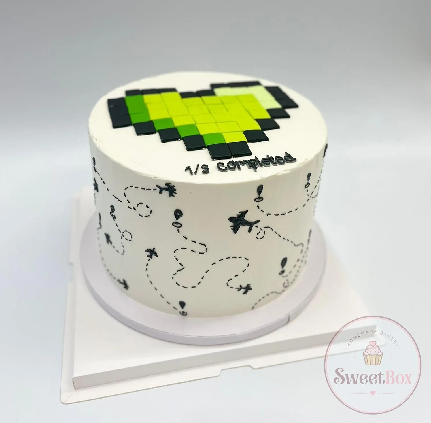 White cake with a green pixel heart made of small squares, 1/3 completed lettering and airplane flight paths on the sides